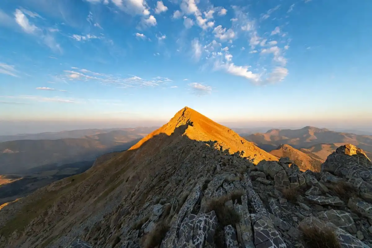 A dramatic mountain peak at sunrise with golden light and rocky terrain in the foreground